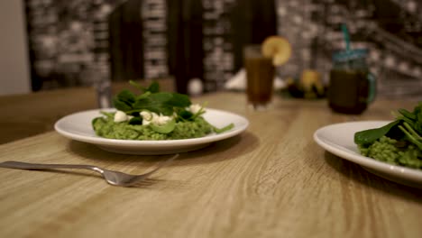 risotto de espinacas verdes servido en dos platos de cerámica blanca sobre una mesa de madera con dos batidos de frutas en el fondo, sartén a la izquierda