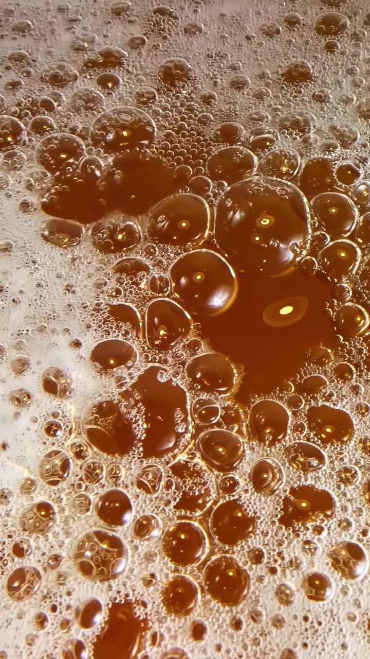 Burbujas doradas suben y estallan en una superficie líquida espumosa, creando un patrón dinámico