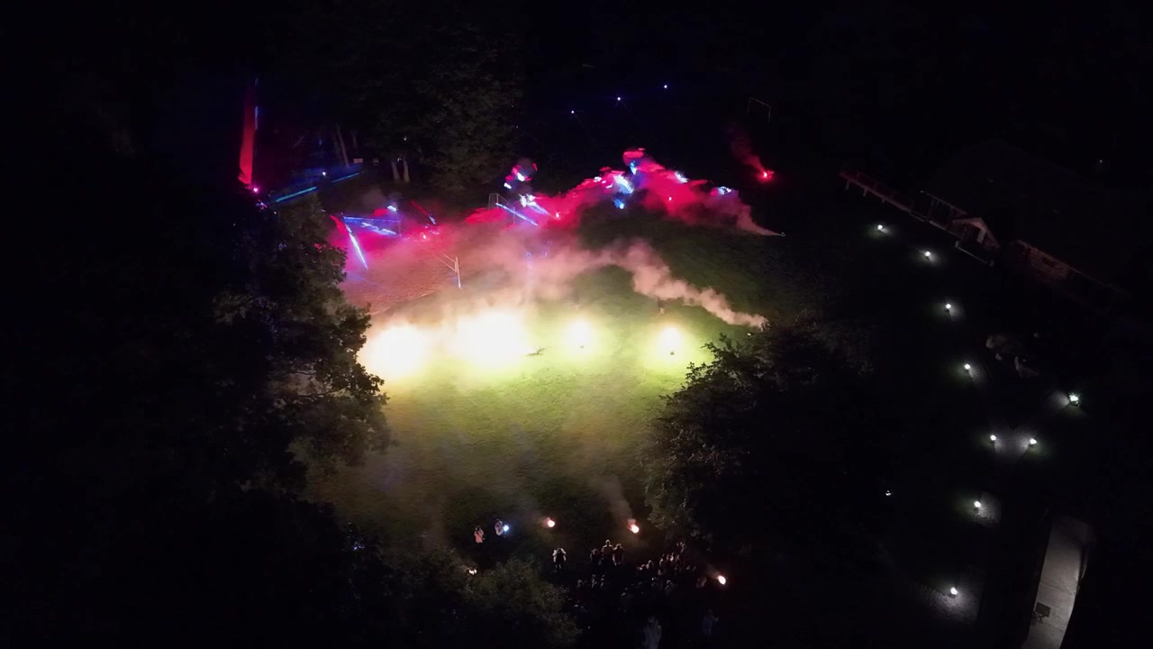 Dronevideo fanger en levende nattebegivenhed udendørs, med levende røgblus, oplyste stier og en folkemængde samlet på et græsareal under dramatisk dæmpet lys