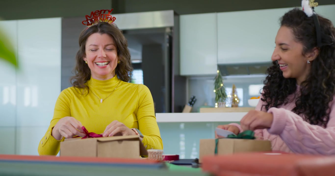 Amiche di diverse etnie che impacchettano regali di carta kraft al tavolo della cucina, legando nastri per le feste.