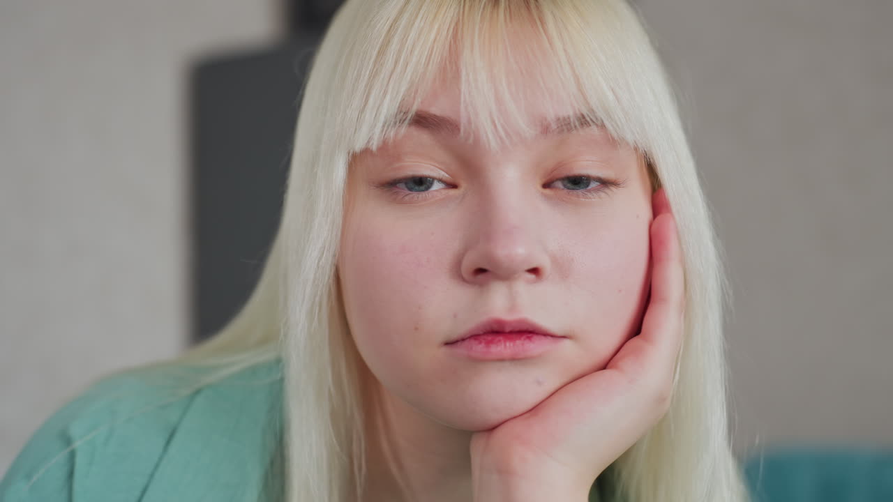 young white woman in green shirt resting head on hand thoughtfully while sitting at kitchen table with decorative plant on table and fridge, microwave in background in modern apartment