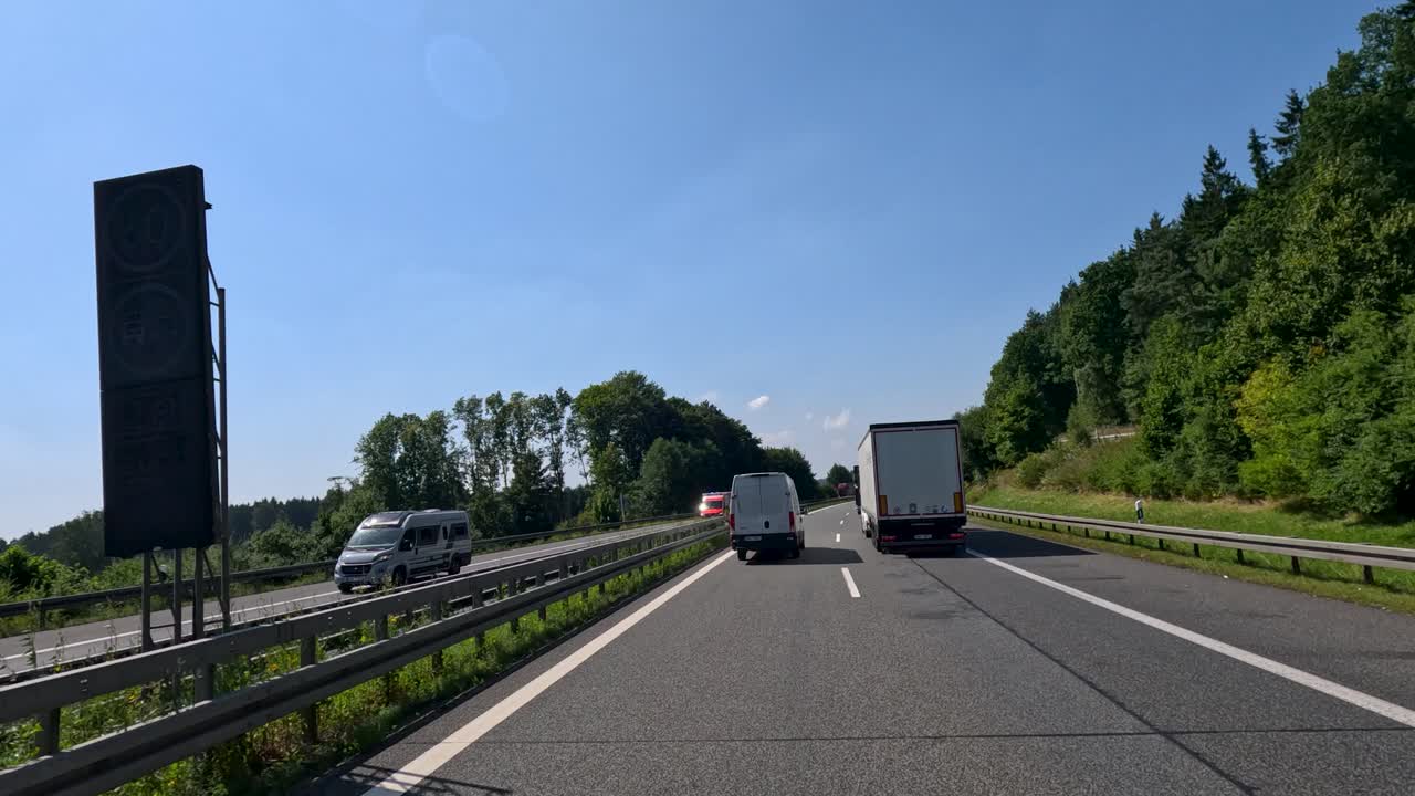 Multiple trucks and vans drive along a sunlit highway, surrounded by greenery, under clear skies