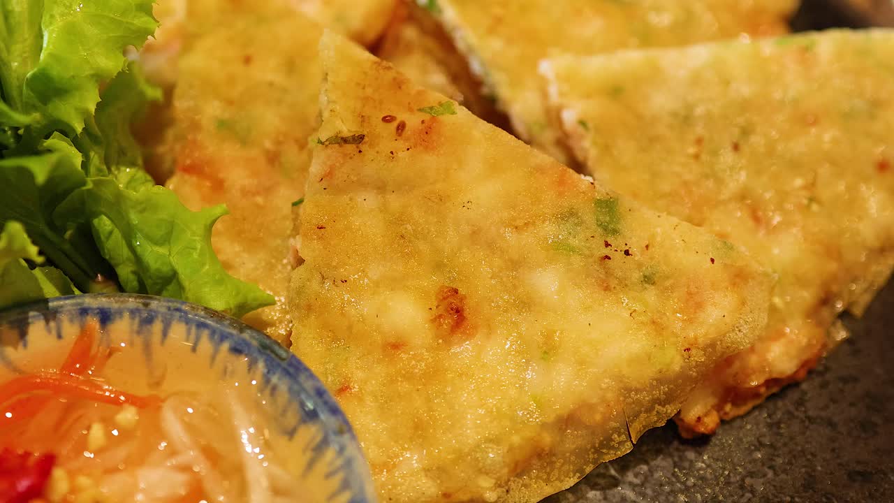 A hand reaches for a Vietnamese pancake on a plate with fresh greens and dipping sauce, under warm lighting