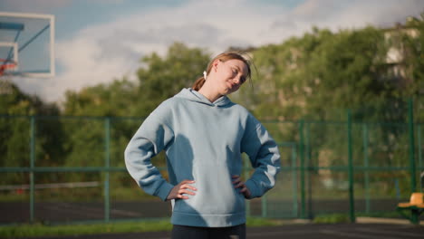 lady in cyan hoodie with hand on waist, working out and turning head from left to right, shadow of another person cast on her with basketball hoop in background