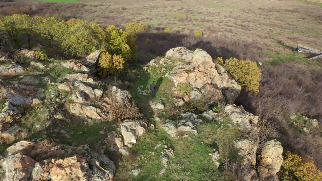 Bird's eye view of man walk to the peak of a mountain, surrounded by large rocks and trees in autumn