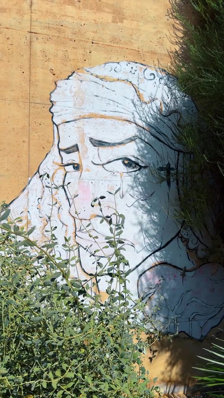 Alicante, Spagna, 8 agosto 2025: Murale di un volto che piange su un muro di cemento. Arte di strada che raffigura un volto in lacrime con un tatuaggio a croce, parzialmente oscurato da cespugli verdi.