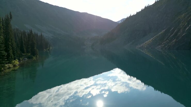 Flying over the lake glassy surface between mountains with morning sun reflecting in the water