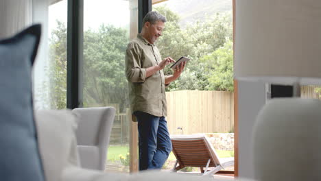 Happy-senior-biracial-man-standing-by-terrace-using-tablet-at-home,-copy-space,-slow-motion