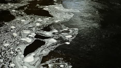 swirling bubbles of white foam slowly float on the surface of dark, black water in a scottish river in a hypnotic, constantly changing pattern where the faster current meets still water