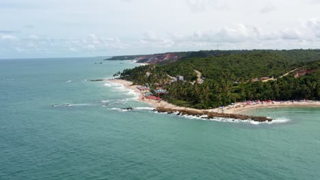 drone aéreo giratorio vista panorámica de la popular playa tropical de coquerinhos cubierta de paraguas con turistas nadando en una piscina natural de un arrecife en condé, paraíba, brasil en un cálido día de verano