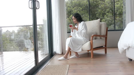 Biracial-woman-wearing-bathrobe-drinking-tea-and-looking-through-window-at-home,-slow-motion