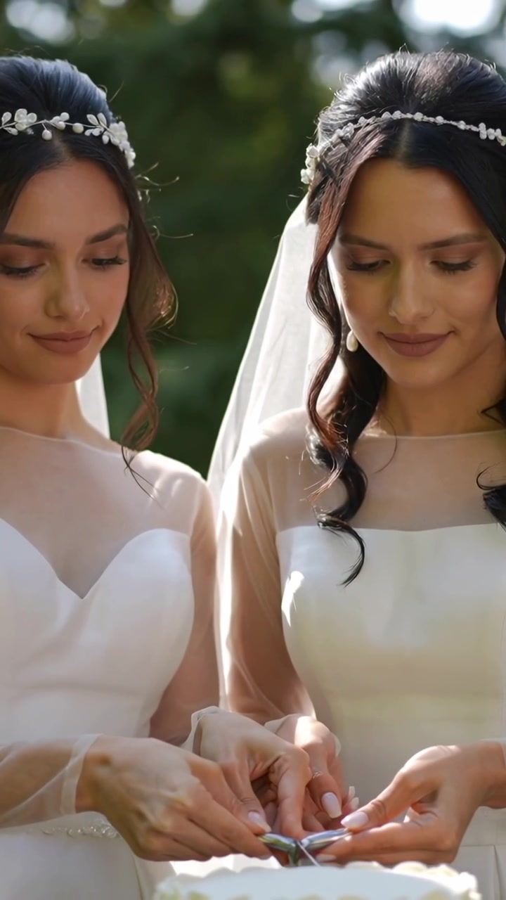 Vidéo verticale : Mariées se tenant la main, coupant le gâteau, toutes deux tenant le couteau dans un parc pour un mariage, en robes.