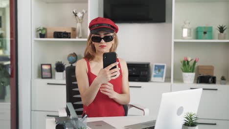 The-young-woman-in-a-red-dress-and-a-red-hat,-wearing-black-glasses,-takes-a-selfie-on-her-phone,-admiring-herself-on-the-camera