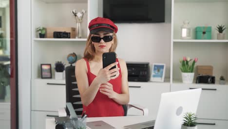 La-Joven-Con-Un-Vestido-Rojo-Y-Un-Sombrero-Rojo,-Con-Gafas-Negras,-Se-Toma-Una-Selfie-En-Su-Teléfono,-Admirándose-A-Sí-Misma-En-La-Cámara