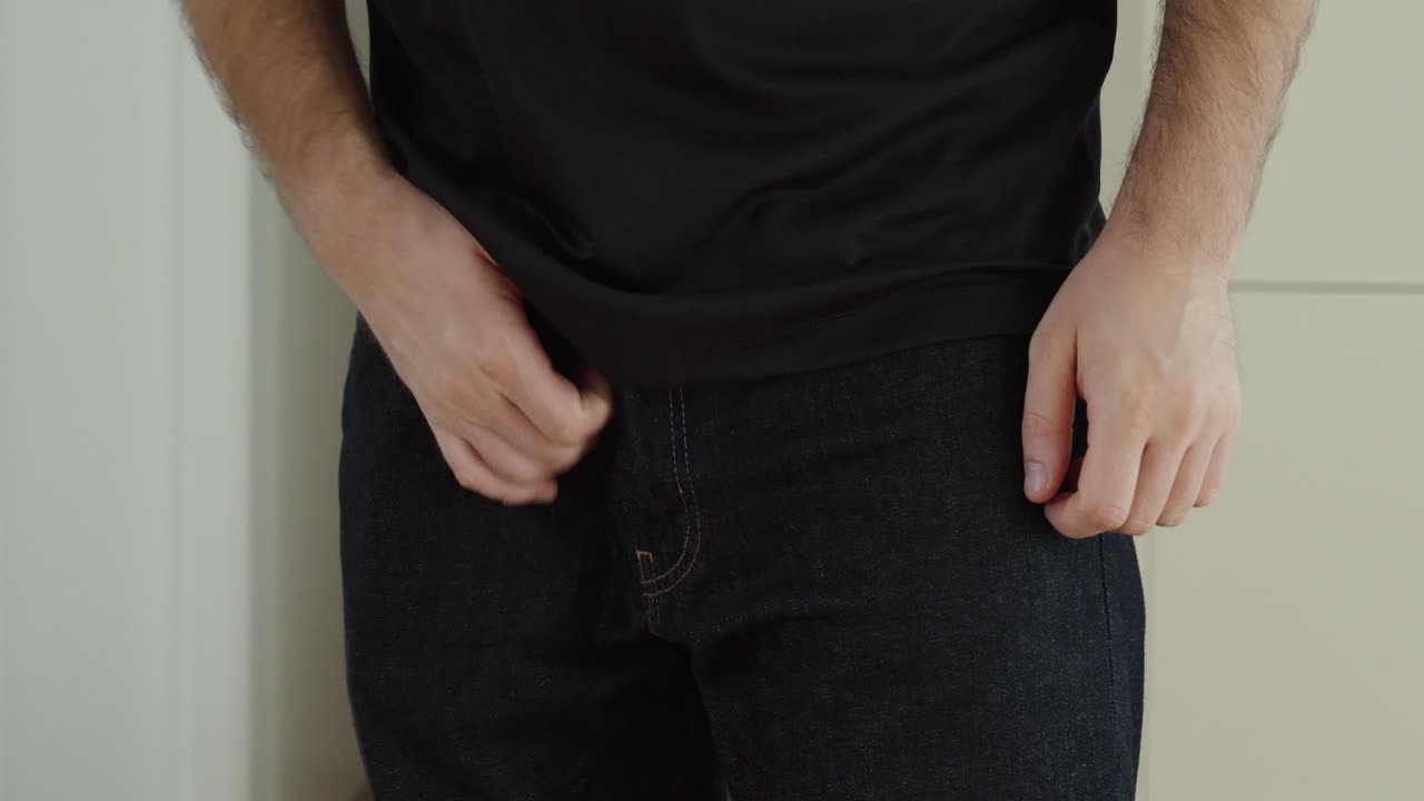 Close-up of a man standing in jeans and adjusting his pants because of itch discomfort, showing personal irritation, awkward body language, and an everyday health-related problem in a realistic indoor