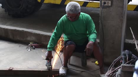 wideshot of an old man with white hair cutting a metal bar using an angle grinder with no protective guards like face shield, gloves, shoes and working suit