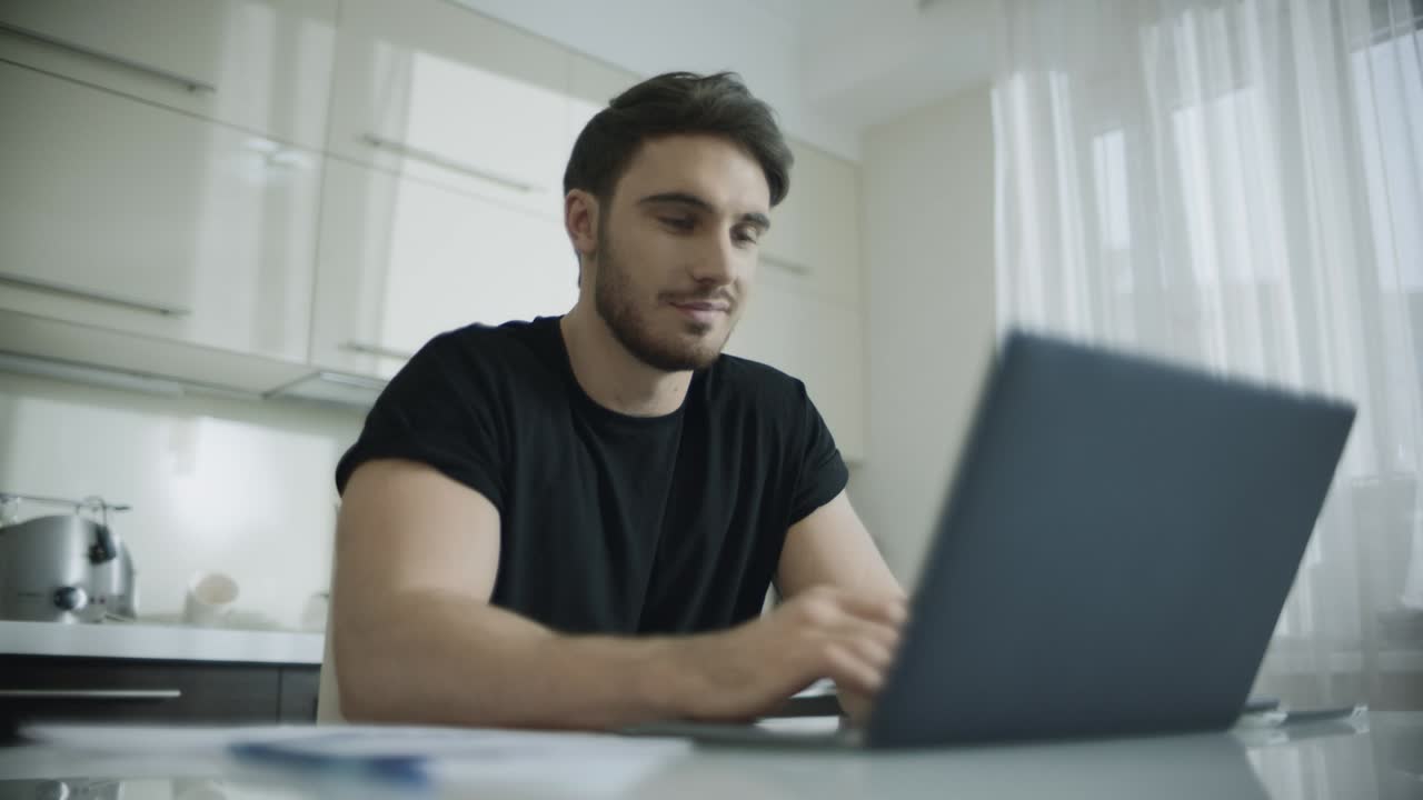 Smiling man using laptop computer at kitchen table. Young business man smiling