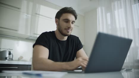 Hombre-Sonriente-Usando-Una-Computadora-Portátil-En-La-Mesa-De-La-Cocina.-Joven-Hombre-De-Negocios-Sonriendo