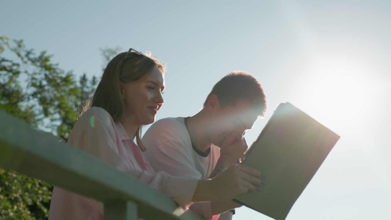 ragazza in top rosa sorridente mentre mostra a suo fratello qualcosa sul suo tablet, lui scuote la testa con una mano e si copre la bocca con l'altra mentre il sole brilla su di loro