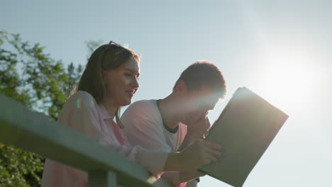 girl in pink top smiling while showing her brother something on her tablet, he shakes his head with one hand and covers his mouth with the other as the sun glows on them