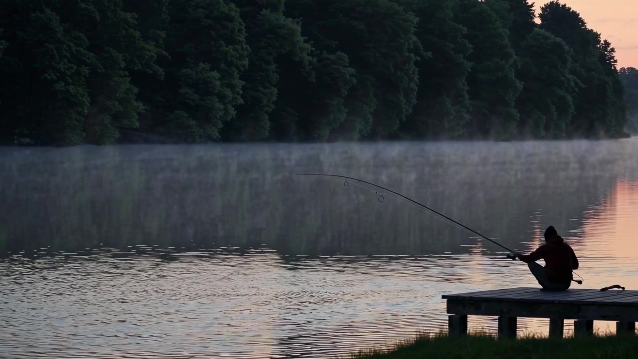 Einsamer Fischer auf einem Steg an einem nebligen See bei Sonnenaufgang