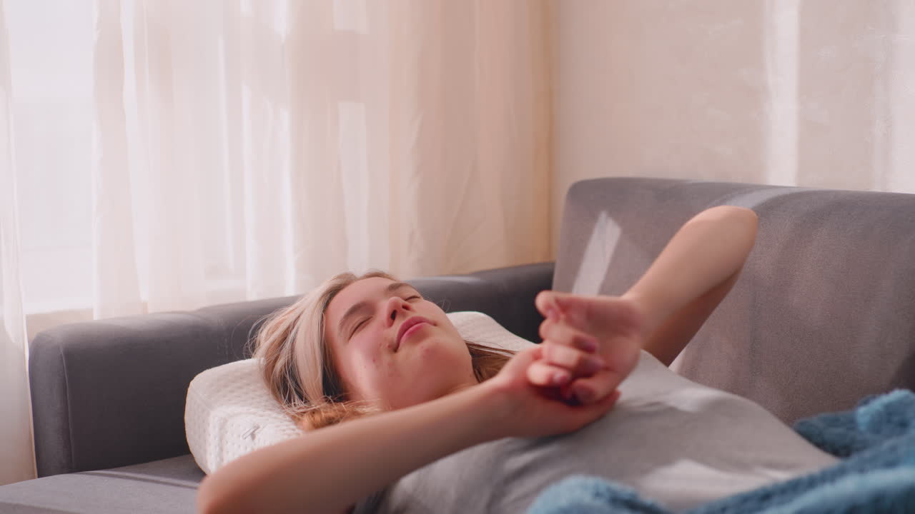 Close up of lady waking up with excitement as she stretches on couch, wrapped in warm blanket, morning sunlight shining through curtains