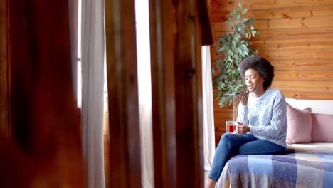 happy african american woman talking on smartphone and drinking tea in living room, in slow motion