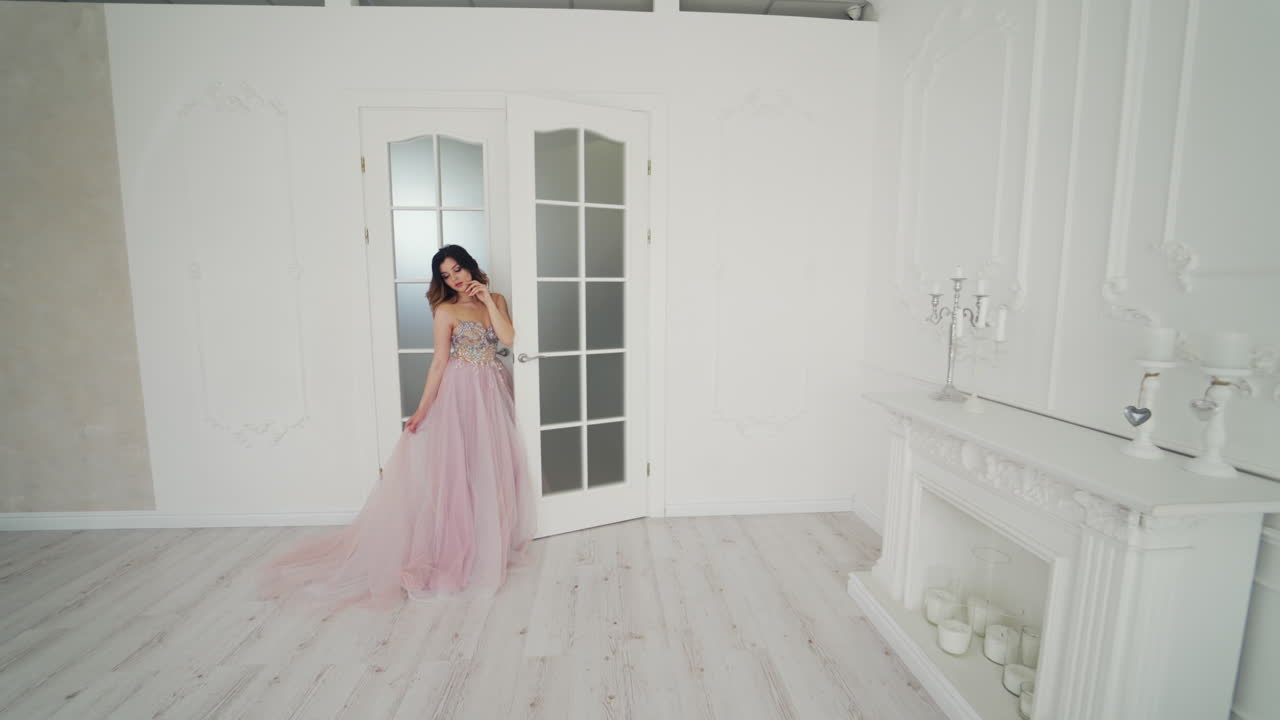 Beautiful bride in wedding dress is standing near the door in the light room. Gorgeous model in luxury dress is posing in studio.