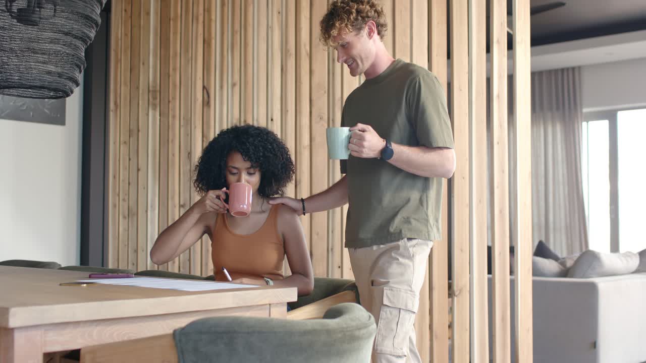 Couple diversifié à la maison, l'homme posant la main sur l'épaule de la femme, la soutenant pendant qu'elle écrit et sirote une tasse rose.