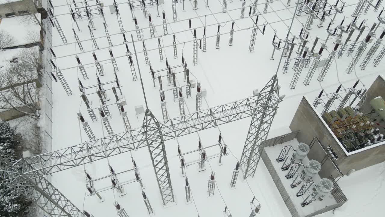 High altitude aerial descent over a snow covered electrical substation, revealing dense arrays of high voltage insulators, steel gantries, and transformers in extreme winter conditions