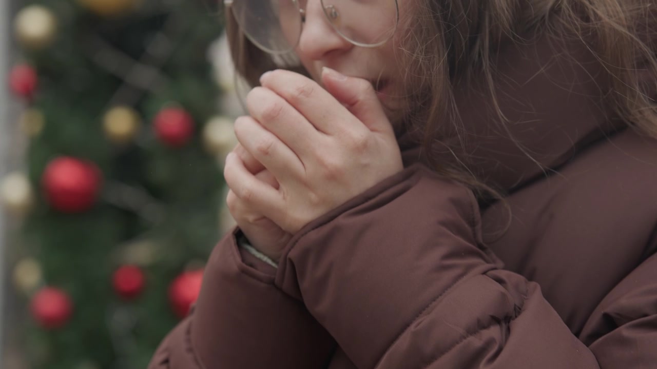 Женщина греет руки у дерева. Сцена холодной погоды с женщиной, греющей руки у праздничного дерева. Молодая женщина в зимней одежде прижимает ладони друг к другу возле украшенного дерева на холодном воздухе.