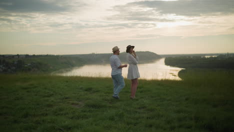 a serene scene where a man in a hat, white shirt, and jeans walks toward a woman in a white dress standing on a grassy hill at sunset, overlooking a peaceful lake