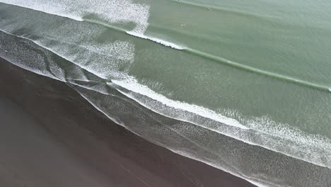 Sets-of-waves-rolling-into-black-sand-beach-with-surfers-waiting-for-a-break-out-the-back