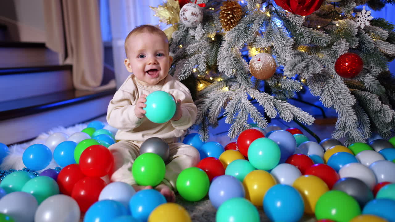 Adorable smiling Caucasian baby sits near the Christmas tree. Happy child plays with the plastic balls scattered around him.