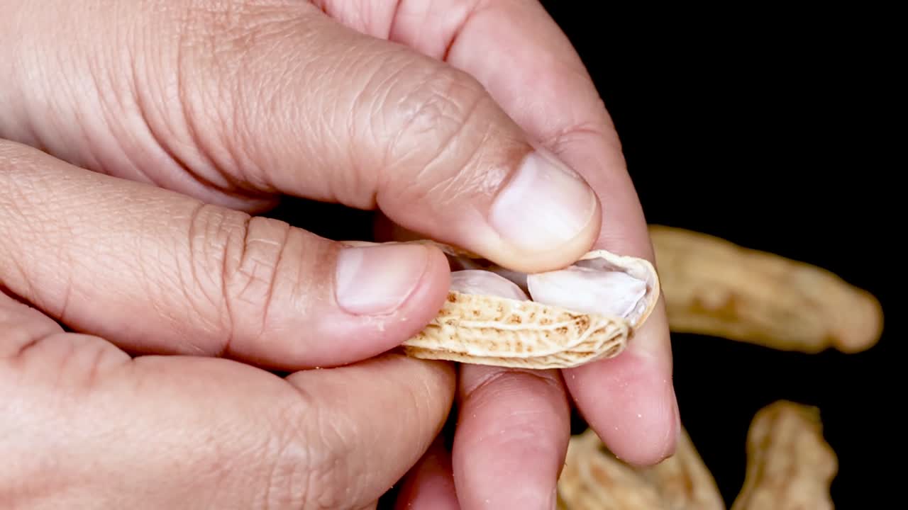 Et detaljeret nærbillede af fingre, der knækker en jordnøddeskal op mod en sort baggrund.