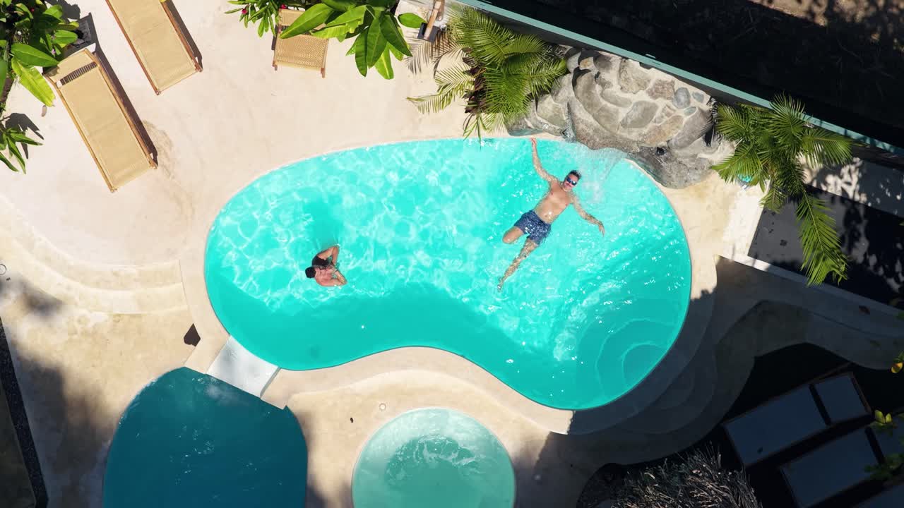 Luftfoto af to personer, der svømmer og flyder i en pool på et tropisk feriested