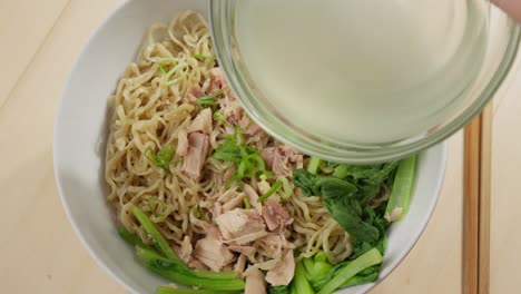 Hand-pours-bowl-of-clear-broth-over-Bakmi-wheat-based-noodles-with-chopsticks-on-side