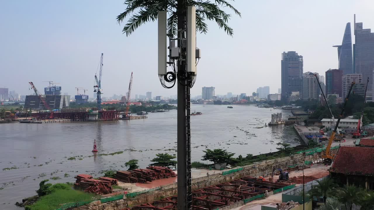 강가에 도시 건물, 크레인, 보트 교통이 있는 강변 건설 현장을 배경으로 야자나무로 위장한 초현대식 휴대전화 탑의 공중 촬영