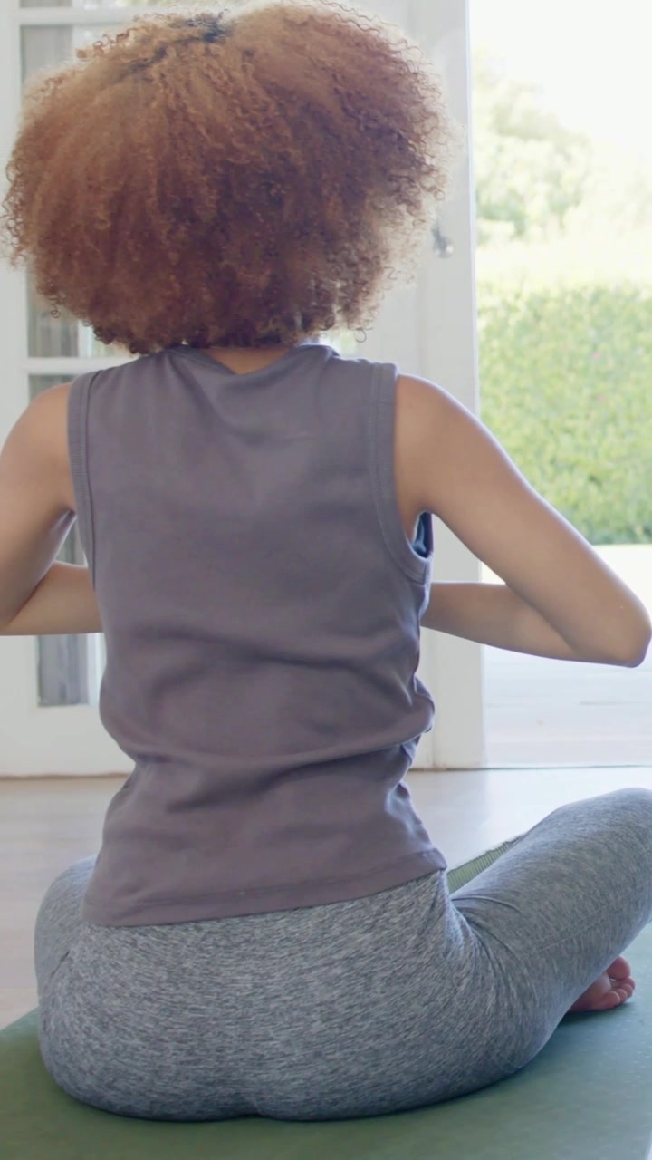 Vertical video: Lowering woman sweeping arms on green mat for yoga by glass doors, tank, smartwatch