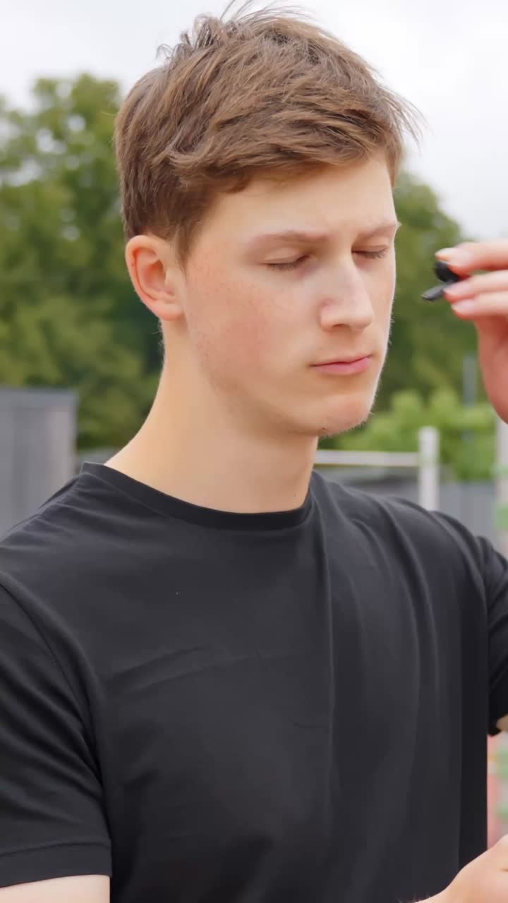 Blue eyed caucasian young man with brown hair, standing in an urban outdoor setting, wearing black t-shirt, adjusts his bluetooth earphones, appearing focused, vertical close up