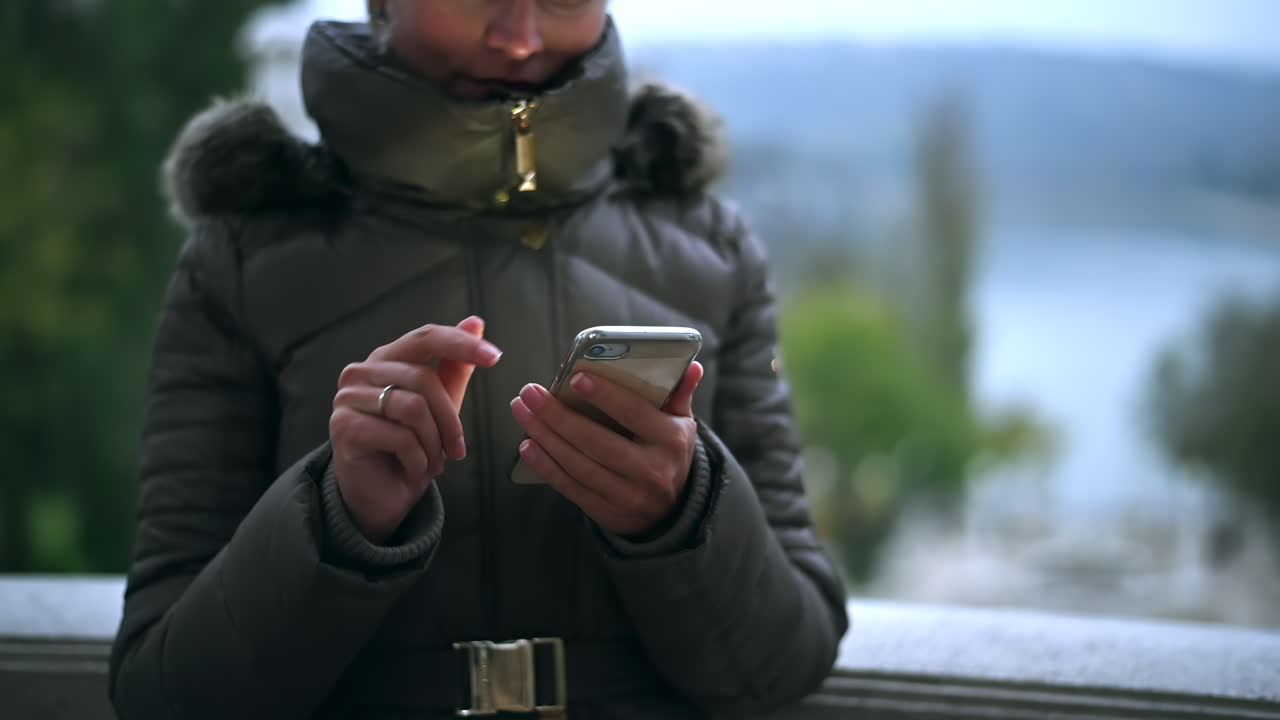 In a tranquil setting by a lake, an individual checks their smartphone, dressed in a warm coat. The cool weather sets a calm mood, surrounded by nature
