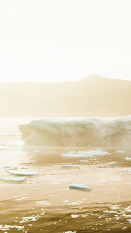 a large iceberg sits on a calm, still ocean with snow and mountains in the background.