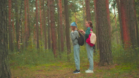 women in serene forest, one wearing teal shirt with red bag, assists her friend in blue bandana with her black bag, tall trees and lush greenery surround them as they prepare for their hike together