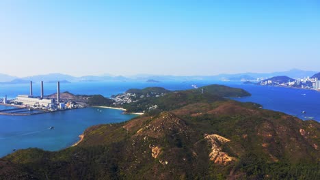 drone shot, traveling forward, over a big green mountain with big coastal coal-fired power plant on an island next to a village with a beach during a beautiful sunny day