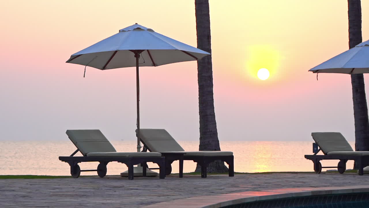Sedie a sdraio sotto gli ombrelloni in un lounge sulla spiaggia al tramonto colorato, riflesso del sole e della luce solare sulla superficie dell'acqua su cielo viola