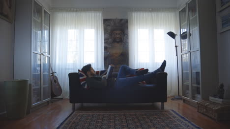 hombre soltero leyendo un libro acostado cómodamente en el sofá en casa frente a la ventana