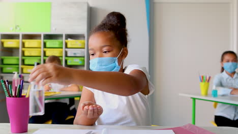 Little-black-girl-with-face-mask-in-class-puts-hand-sanitizer-on-her-hands.-COVID-19-pandemic.-Children-back-to-school-after-coronavirus-lockdown.