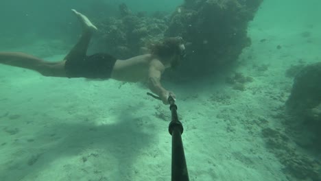 un hombre joven, en forma y fuerte con cabello largo y barba está nadando bajo el agua en un agua turquesa vibrante con arrecifes de coral a su lado y burbujas girando de sus movimientos
