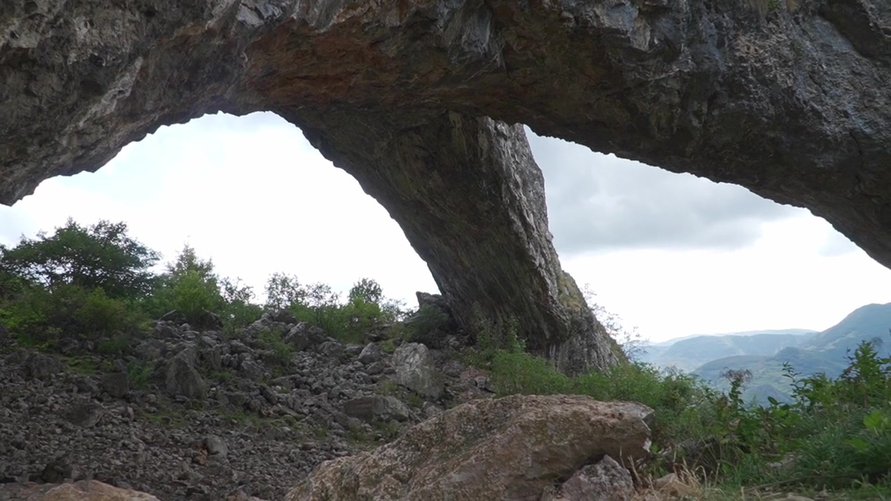 En naturlig steinbue rammer inn utsikten over ulendt terreng og fjerne fjell under en skyet himmel