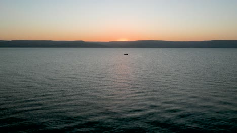 Aerial-drone-view,-slow-movement:-flying-over-the-sea-at-sunrise-or-sunset,-with-a-lone-boat-in-the-background,-an-orange-horizon-line-and-calm-waves-creating-a-relaxing-pattern